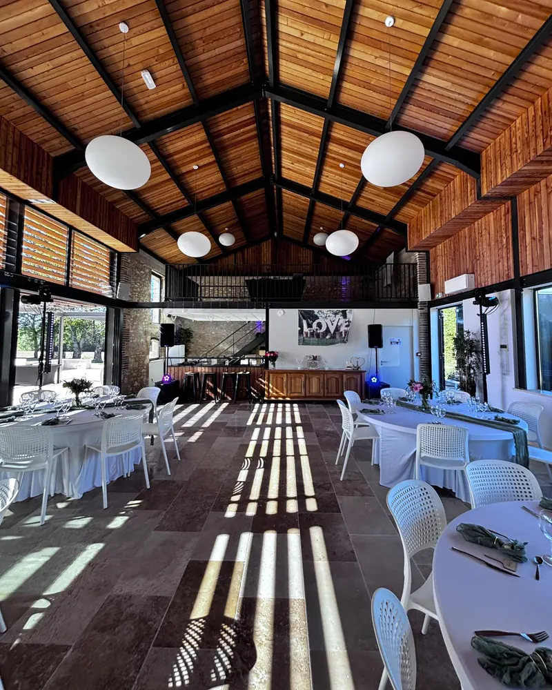 Salle de réception de mariage sous une charpente en bois : suspensions blanches, tables rondes nappées de blanc, serviettes vertes.